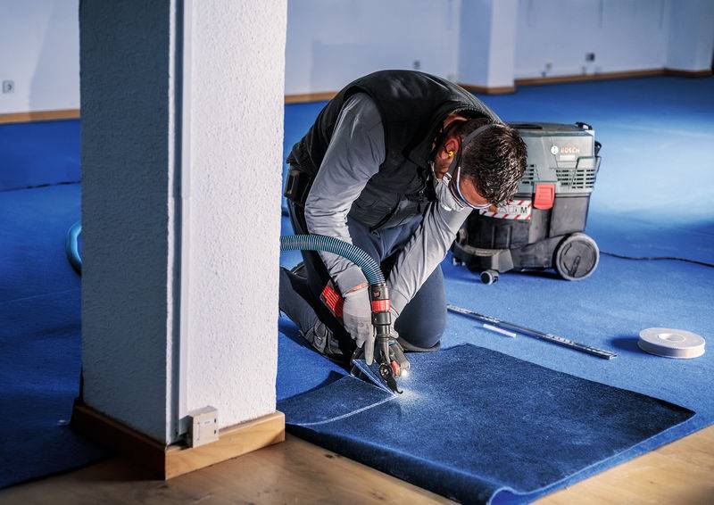 Ein Handwerker schneidet Teppichboden in einem Raum mit blauer Auslegware. Neben ihm stehen ein Staubsauger und Werkzeuge.