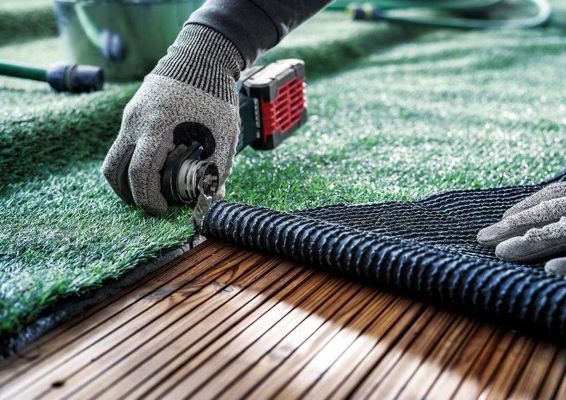 Eine Person schneidet Kunstrasen mit einem Messer auf einer Holzterrasse zu. Die Hände tragen Arbeitshandschuhe.