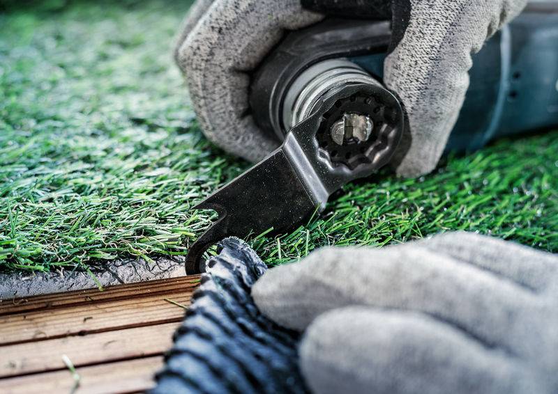 Handwerker verwendet ein Messer, um Kunstrasen auf eine Holzoberfläche zuzuschneiden. Handschuhe schützen die Hände während der Arbeit.