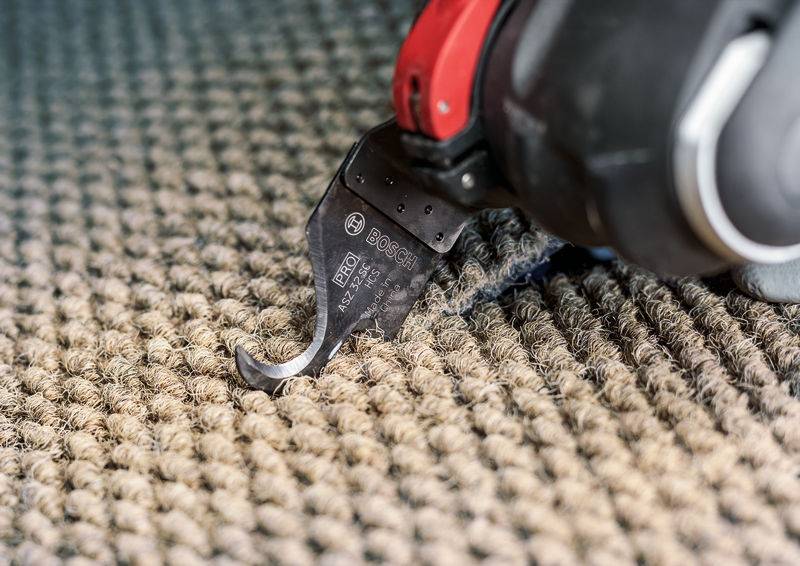 Ein elektrisches Schneidwerkzeug schneidet Teppich auf. Das Bild zeigt den scharfen, gebogenen Aufsatz des Werkzeugs über der Teppichoberfläche.