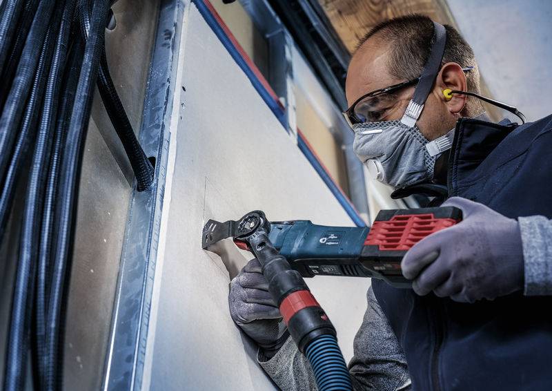 Eine Person trägt Schutzkleidung und schneidet mit einem Elektrowerkzeug ein Stück aus einer Wand. Dabei trägt sie Gehörschutz und eine Schutzbrille.