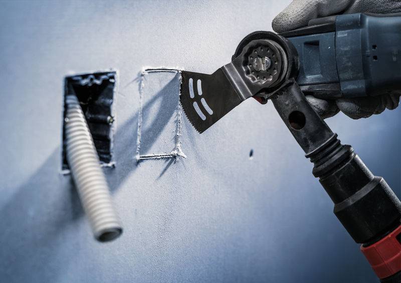 Eine Hand mit Handschuh hält ein oszillierendes Multitool, das ein rechteckiges Loch in eine graue Wand schneidet. Ein Rohr ragt aus einem weiteren Loch hervor.