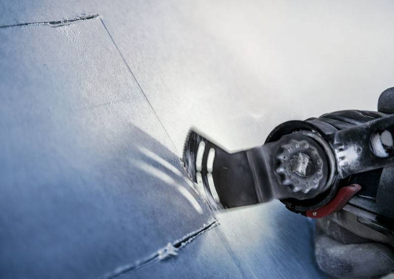 Eine Hand hält ein oszillierendes Werkzeug, das eine Metalloberfläche schneidet oder schleift. Werkstück und Werkzeug sind im Fokus.