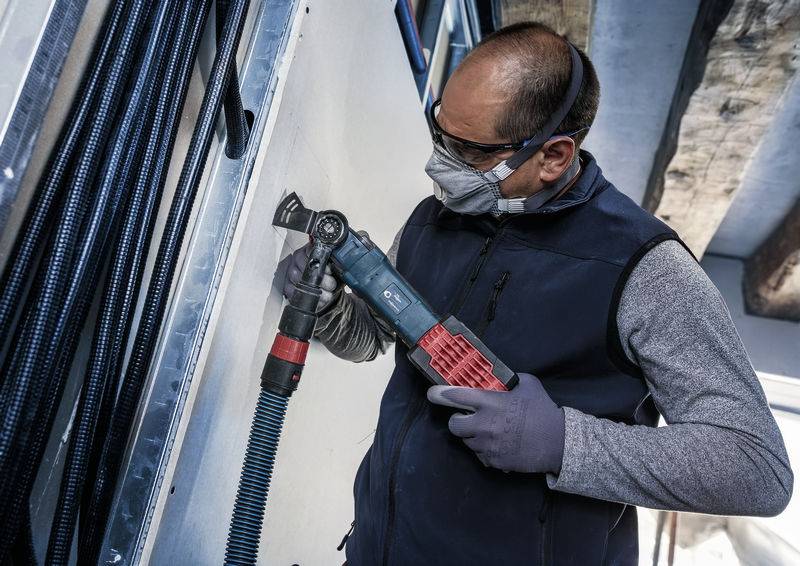 Eine Person, die Schutzbrille und Maske trägt, schneidet mit einem Elektrowerkzeug durch eine Wand. Im Hintergrund sind Kabel sichtbar.