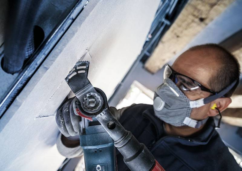 Ein Arbeiter trägt Schutzbrille und Atemmaske und schneidet mit einem Elektrowerkzeug in eine Wand.