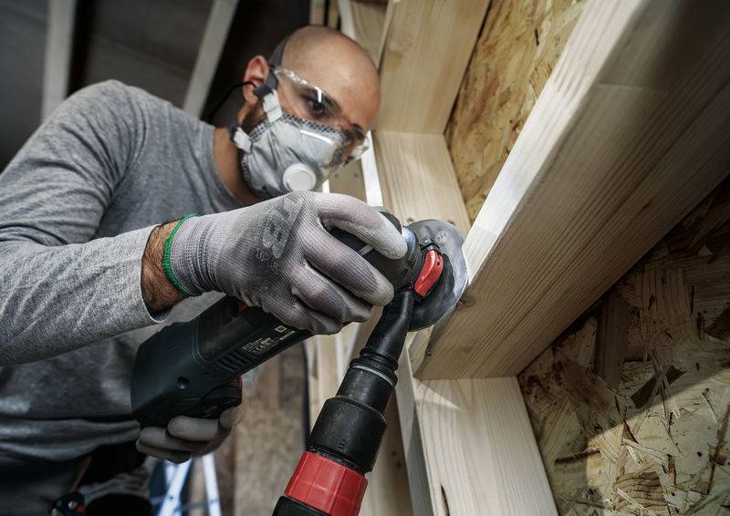 Ein Handwerker schleift Holz mit einem Elektrowerkzeug. Er trägt Schutzbrille und Atemmaske in einer Werkstattumgebung.