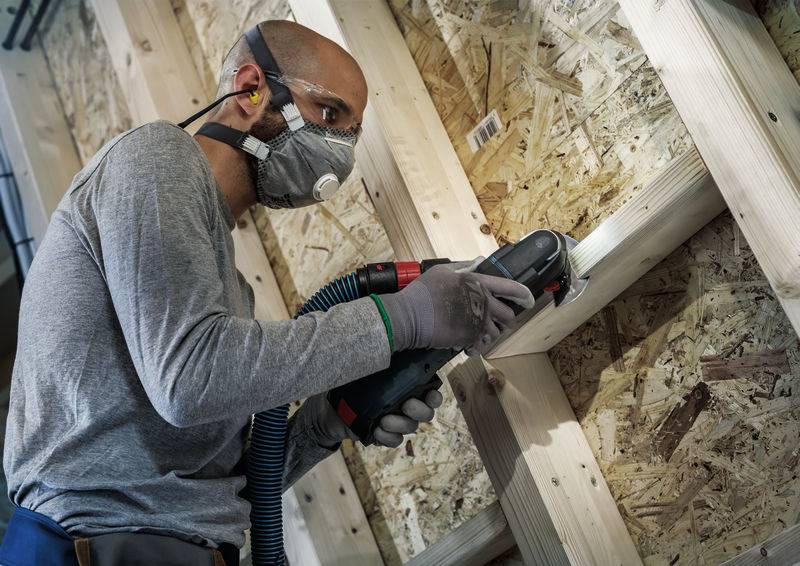 Ein Mann trägt Schutzkleidung und schleift Holz mit einem Elektrowerkzeug in einer Werkstatt.