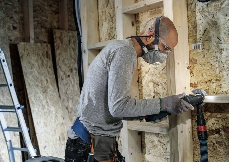 Ein Bauarbeiter mit Schutzbrille und Maske schleift Holz in einer Werkstatt. Eine Leiter und Holzplatten sind im Hintergrund zu sehen.