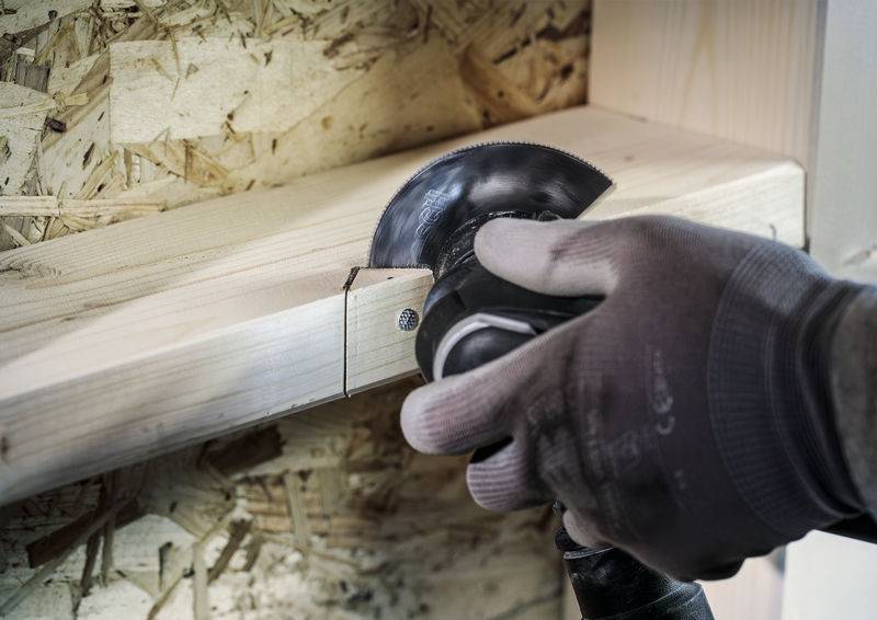 Hand sägt Holzbrett in Werkstattumgebung. Hintergrund aus grobem Holz und Sperrholz. Person trägt Arbeitshandschuhe.