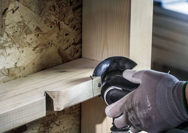 Person schleift Holz mit einem Elektrowerkzeug. Schutzbrille und Handschuhe werden getragen.
