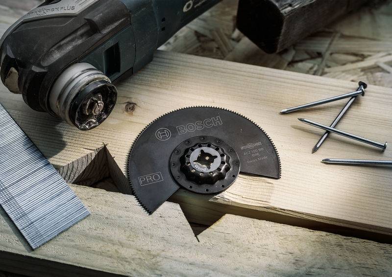 Ein multifunktionales Oszillationswerkzeug mit Sägeblatt liegt auf einem Holzbrett, daneben einige Nägel und Metallklammern.