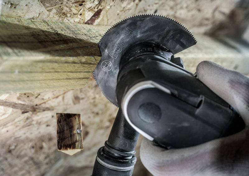 Eine Person schneidet mit einem oszillierenden Werkzeug ein Stück Holz, das an eine Holzwand genagelt ist.