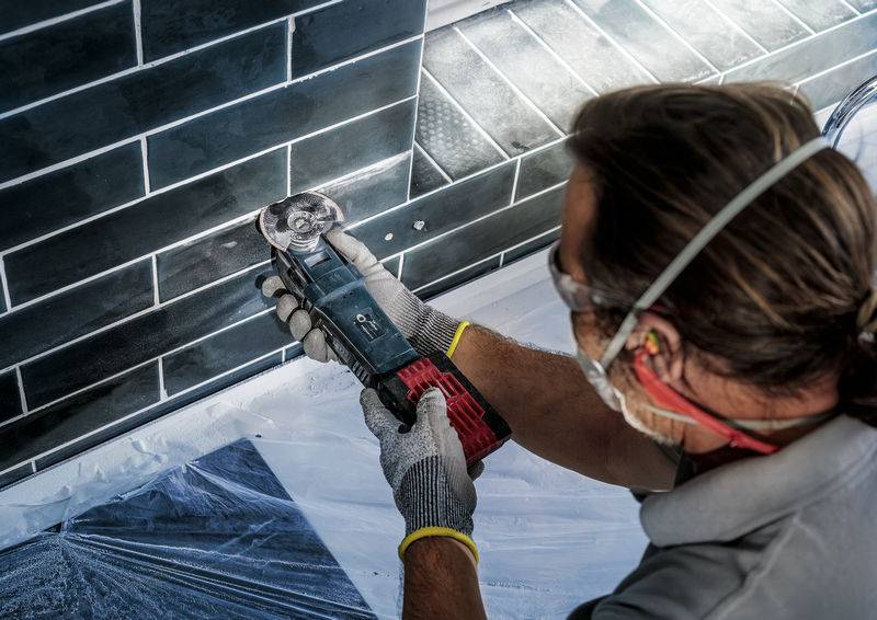 Eine Person schneidet präzise schwarze Fliesen mit einem elektrischen Werkzeug. Schutzbrille und Handschuhe werden getragen.