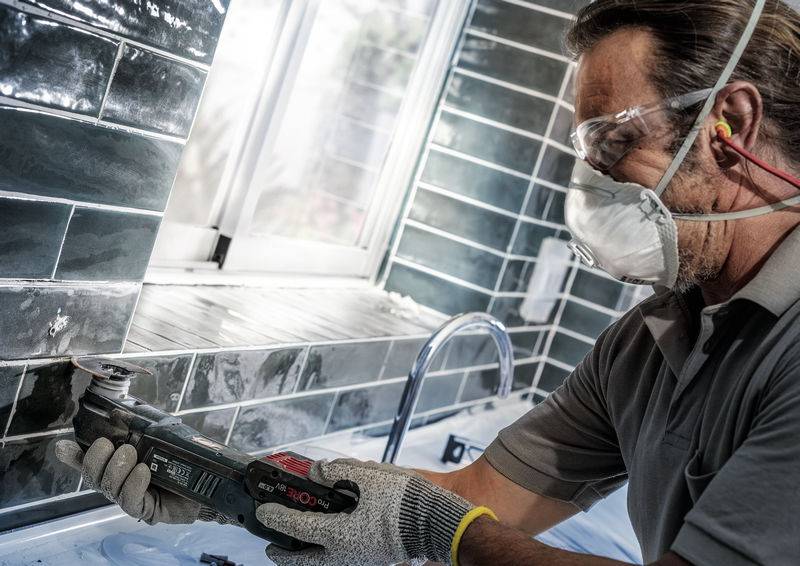 Ein Handwerker mit Schutzbrille und Maske verwendet ein Elektrowerkzeug, um Fliesen in einem Badezimmer zu schneiden.