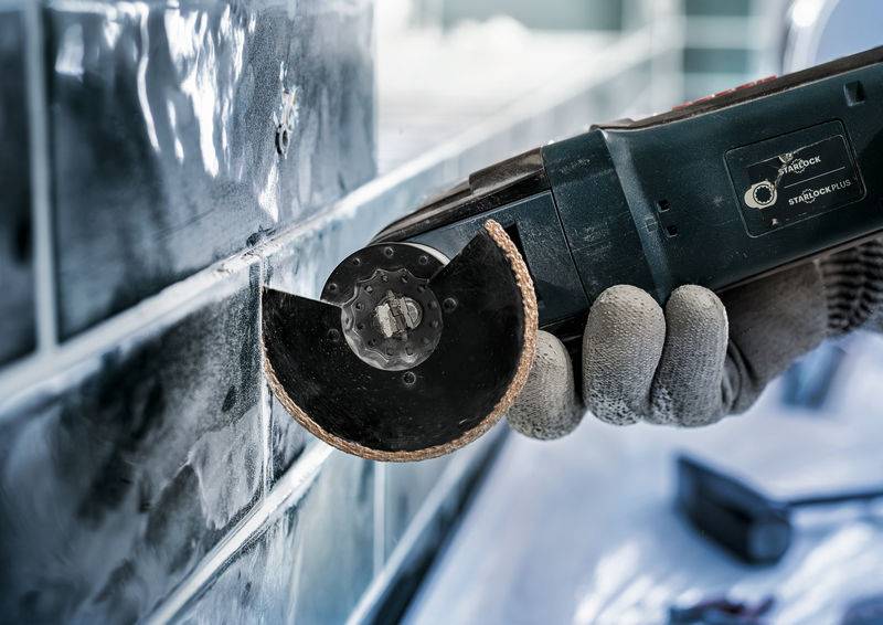 Eine Person benutzt ein elektrisches Trennwerkzeug mit einer Sägeklinge, um Fugen zwischen schwarzen Fliesen zu schneiden. Arbeitsumgebung sichtbar.