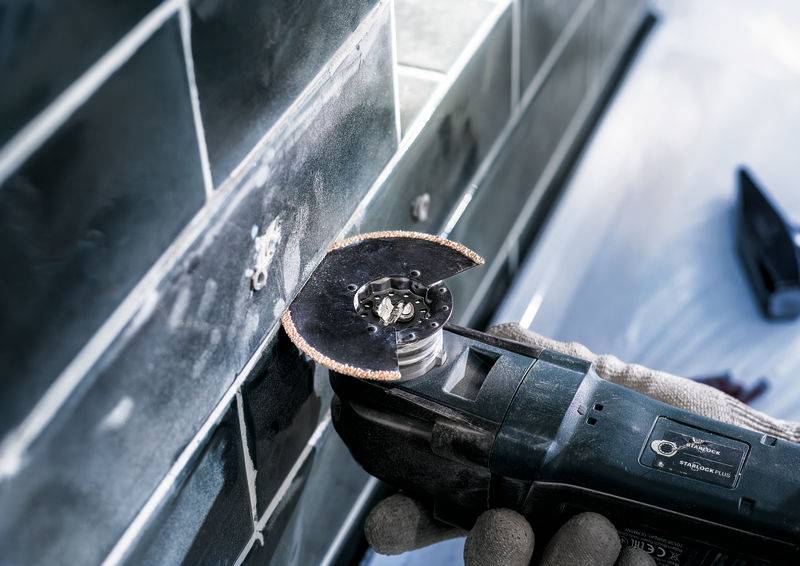 Hand hält Winkelschleifer, der Fliesen an einer Wand schneidet. Trägt Schutzhandschuhe. Arbeitet im Innenraum mit glänzenden Fliesen.