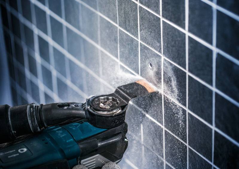 Eine Person verwendet ein Elektrowerkzeug, um ein quadratisches Fliesenloch in eine Wand zu schneiden. Staub ist sichtbar.