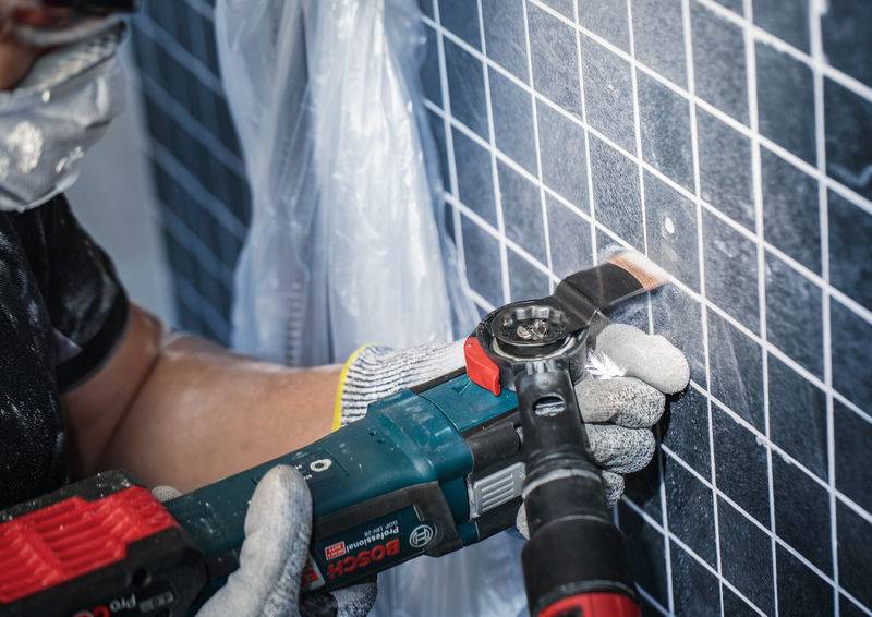 Eine Person benutzt ein Elektrowerkzeug, um Fliesen an einer Wand zu bearbeiten. Schutzbrille und Handschuhe werden getragen.