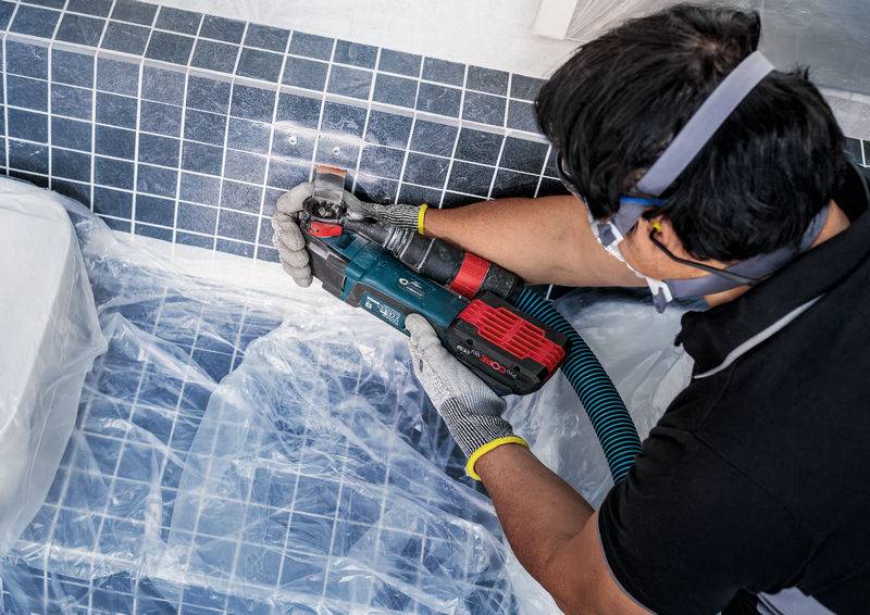 Eine Person bohrt mit einem Elektrowerkzeug in eine geflieste Wand. Schutzkleidung und Staubschutz sind vorhanden.