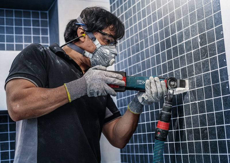 Eine Person trägt Schutzkleidung und verwendet ein elektrisches Werkzeug, um Fliesen an einer Wand zu bearbeiten.