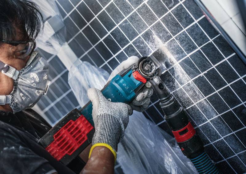 Person mit Schutzbrille und Maske schneidet mit Elektrowerkzeug in eine Solarpanel-Oberfläche. Arbeitsplatz mit Plastikhülle abgedeckt.
