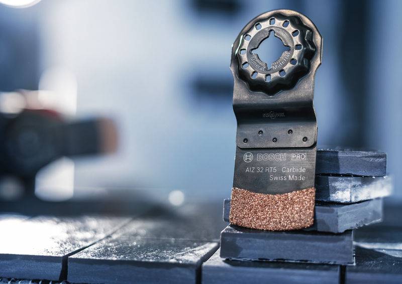 Ein oszillierendes Sägeblatt steht schräg auf gestapelten Fliesen. Es trägt die Aufschrift 'Carbide Swiss Made'.