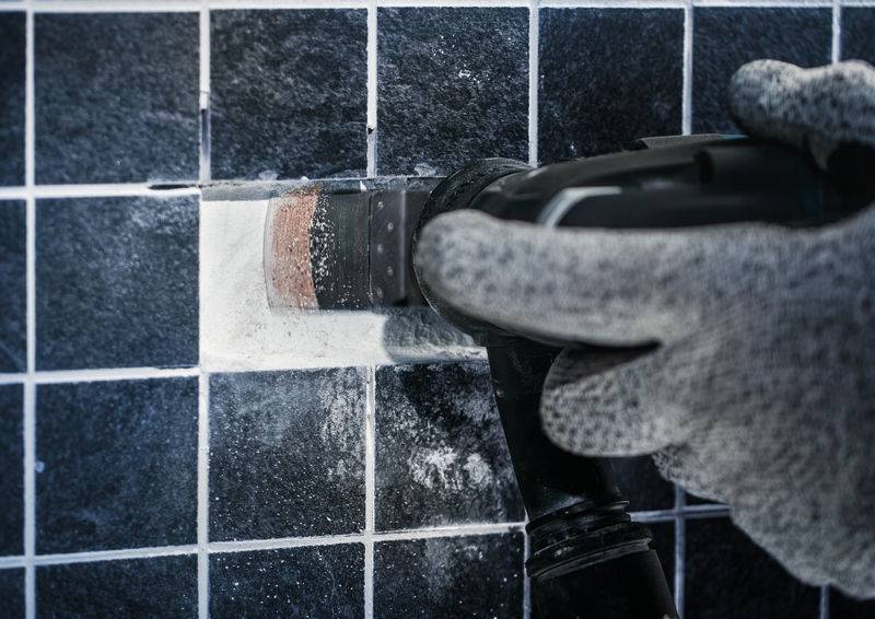 Hand in grauem Handschuh benutzt ein elektrisches Werkzeug, um Fugenmörtel zwischen dunklen Fliesen zu entfernen.