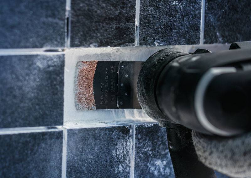 Ein Werkzeug entfernt Fliesenfugen von einer dunklen Kachelwand. Eine Schneidescheibe bearbeitet die weiße Fugenmasse.