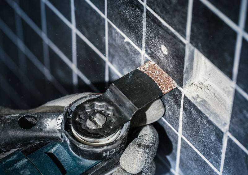 Eine Hand mit Handschuh hält ein oszillierendes Werkzeug, das eine Fuge zwischen dunkelgrauen Kacheln bearbeitet. Staub entsteht.