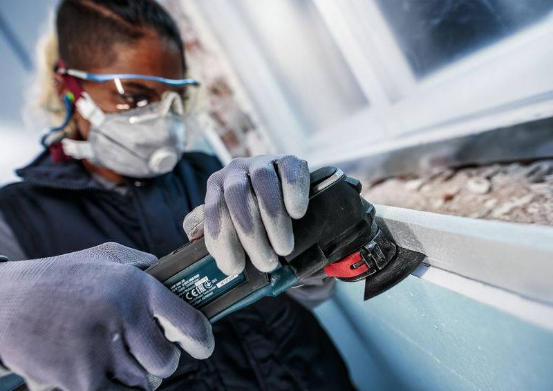 Eine Person trägt Schutzbrille, Maske und Handschuhe und arbeitet mit einem Winkelschleifer an einer Oberfläche.