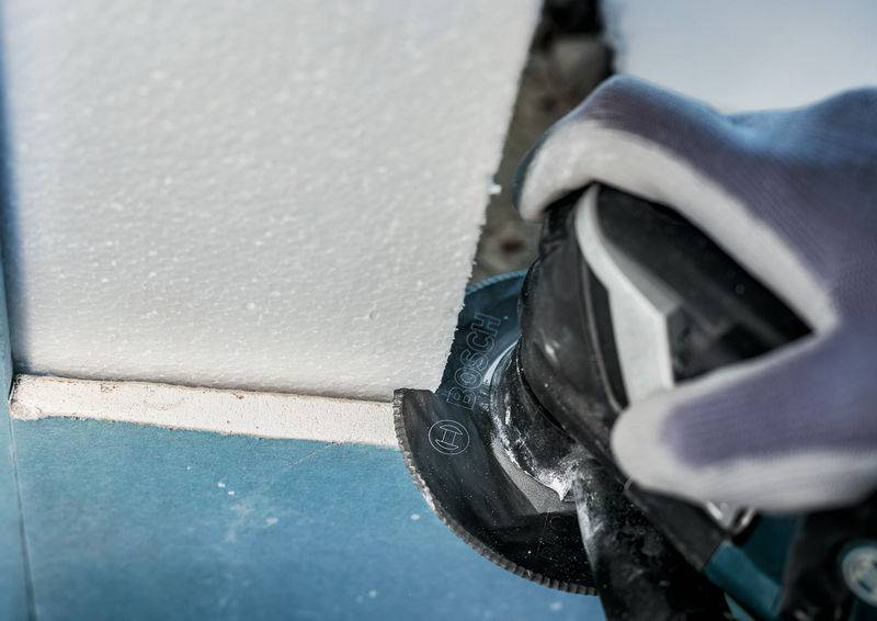 Hand mit Handschuh hält ein Elektrowerkzeug, das Styropor schneidet. Nahaufnahme zeigt präzises Arbeiten an einer Wandkante.