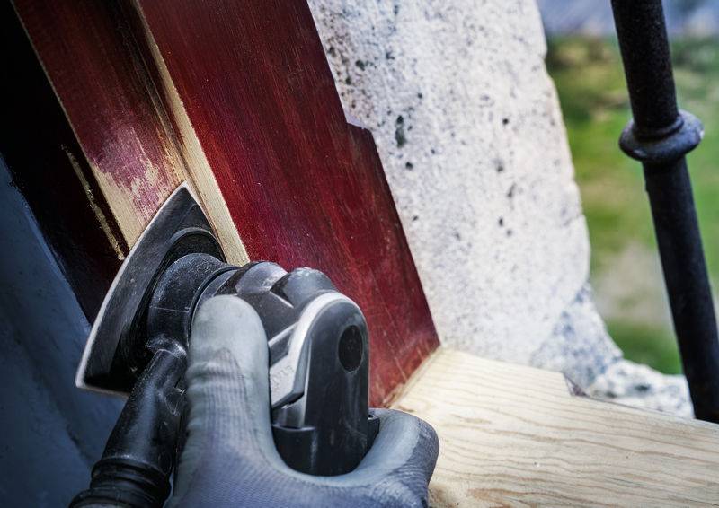 Eine Person schleift Holz an einem Fensterrahmen mit einer elektrischen Schleifmaschine, um es für die Lackierung vorzubereiten.