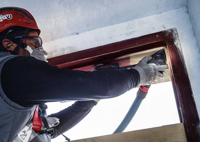 Eine Person in Schutzkleidung nutzt ein Elektrowerkzeug, um an einem Türrahmen zu arbeiten, möglicherweise zur Renovierung oder Reparatur.