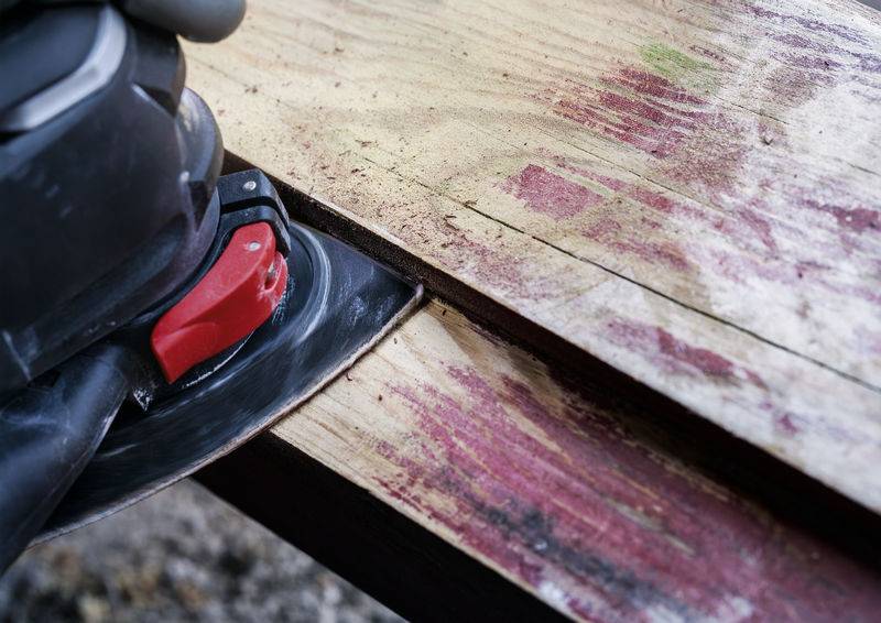 Eine Schleifmaschine glättet die raue Holzoberfläche eines Tisches. Rote Rückstände sind sichtbar, was auf abgeschliffene Farbe hinweist.