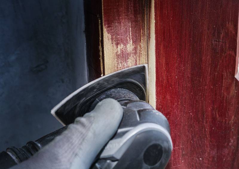 Nahaufnahme einer Person, die mit einem Schleifgerät roten Lack von einer Holzoberfläche entfernt. Eine Schutzhandschuh ist sichtbar.