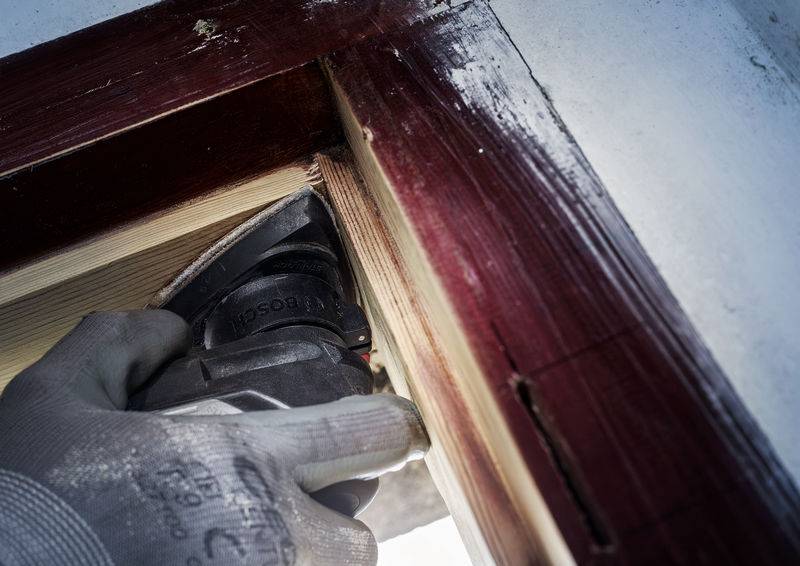 Eine Hand mit einem Handschuh hält ein Elektrowerkzeug und schneidet Holz an einem rot lackierten Fensterrahmen.