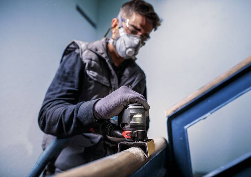 Eine Person schleift mit einer elektrischen Schleifmaschine ein Holzgeländer. Sie trägt eine Schutzbrille und eine Atemschutzmaske.