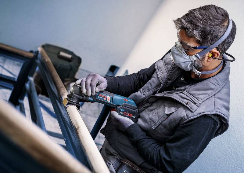 Ein Mann bearbeitet mithilfe eines Elektrowerkzeugs ein Geländer. Er trägt Sicherheitsausrüstung, einschließlich Schutzbrille und Gehörschutz.