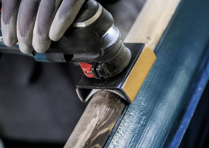 Eine Person schleift mit einem Schleifgerät die Kante eines Holzstücks. Auf der linken Seite ist ein Handschuh zu sehen.