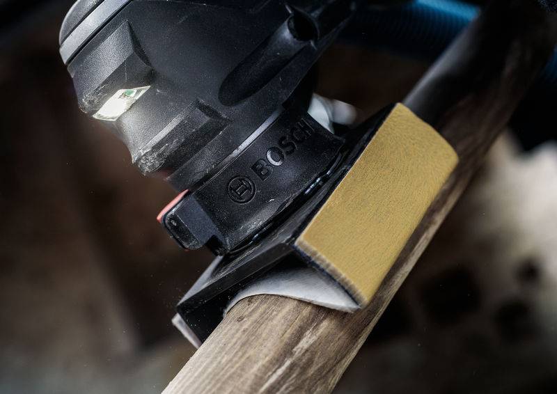 Ein Schwingschleifer bearbeitet eine Holzkante, erkennbar an der markanten Schleiffläche und der Staubschutzvorrichtung im Einsatz.