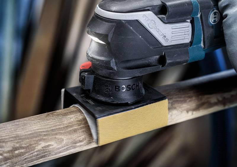 Eine Person verwendet ein elektrisches Bosch-Werkzeug zum Schleifen eines Holzbalkens. Das Gerät hat eine rechteckige Schleiffläche.