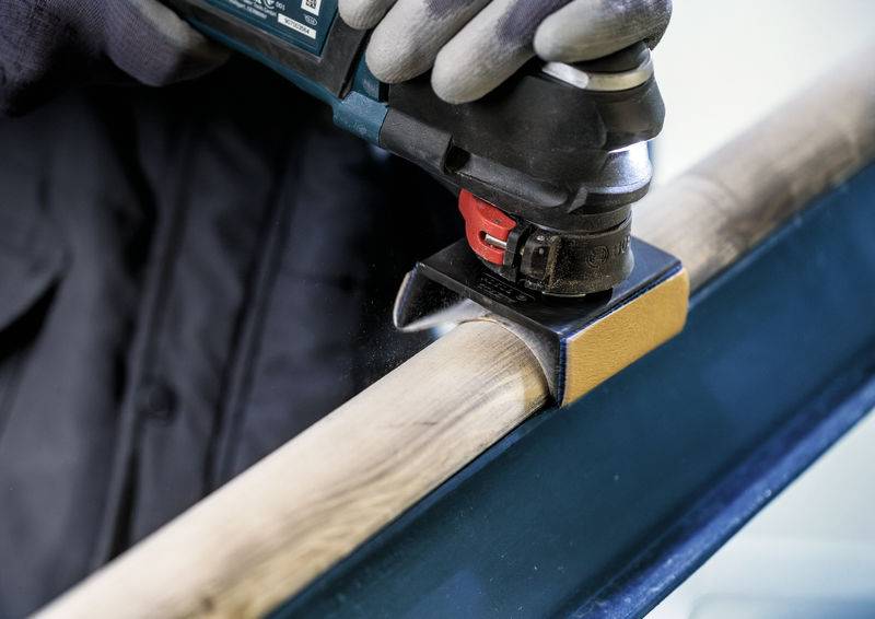 Eine Person schleift mit einem Elektrowerkzeug eine Holzstange. Sie trägt Arbeitshandschuhe. Das Werkzeug erzeugt Staub.