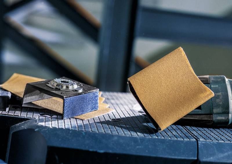 Ein Tisch mit technischem Gerät und Schleifpapier. Im Hintergrund sind unscharfe Metallstrukturen sichtbar.