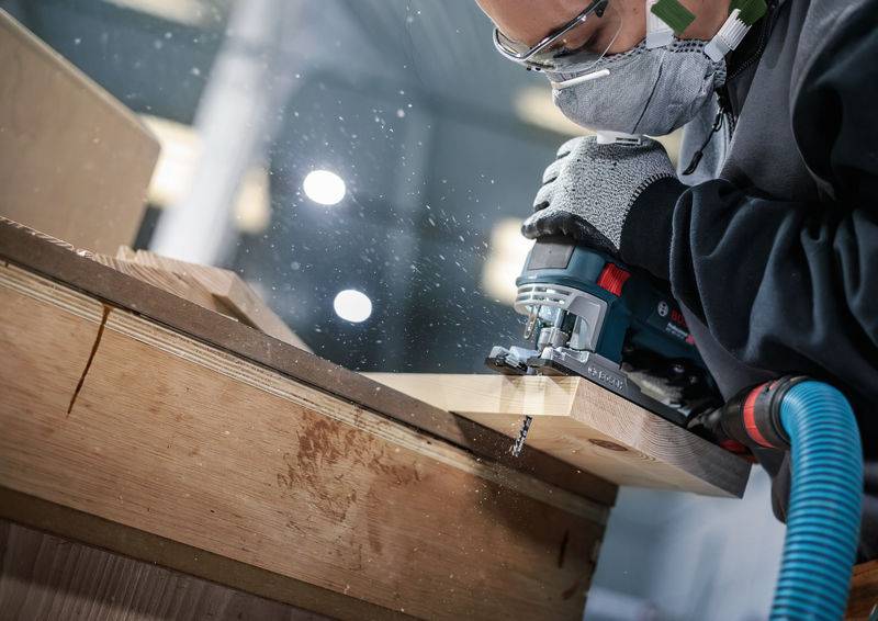 Eine Person mit Schutzbrille und Maske verwendet ein Elektrowerkzeug, um ein Holzbrett in einer Werkstatt zu schneiden.