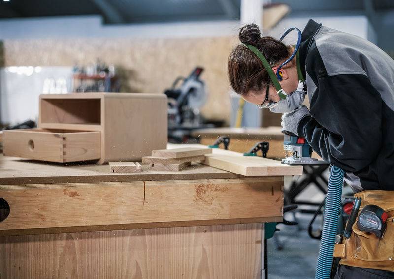 Eine Person trägt Schutzbrille und Gehörschutz, während sie Holz an einem Arbeitstisch in einer Schreinerwerkstatt bearbeitet.