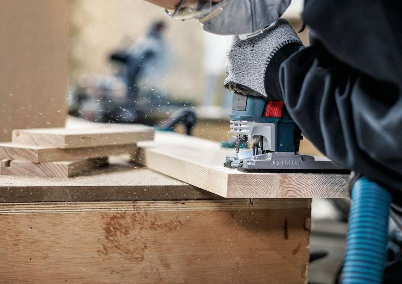 Person arbeitet mit einer Oberfräse an einem Holzbrett; Späne fliegen durch die Luft. Schutzhandschuhe und Werkzeug sind sichtbar.