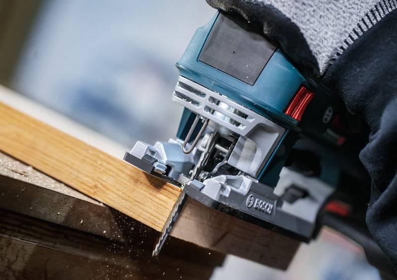 Eine elektrische Stichsäge durchschneidet ein Holzbrett. Sägespäne fliegen in die Luft. Arbeitshandschuhe schützen die Hände.