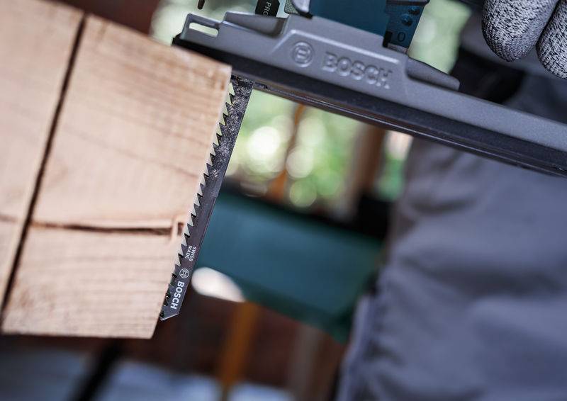 Nahaufnahme einer Säge, die durch ein Holzbrett schneidet. Ein Handschuh hält die Säge stabil. Bosch-Logo ist erkennbar.