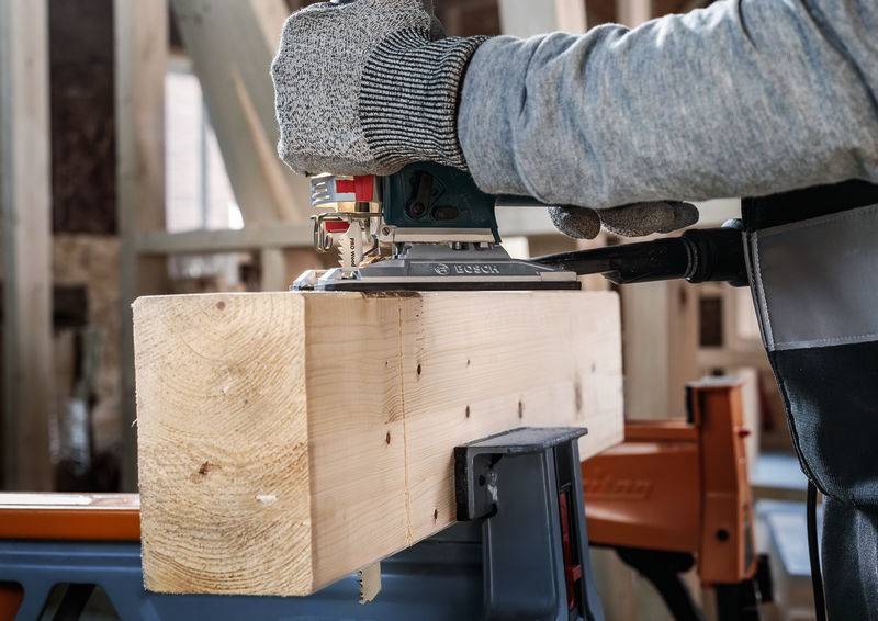 Eine Person hobelt einen Holzblock mit einer elektrischen Hobelmaschine. Sie trägt Handschuhe zum Schutz bei der Arbeit.