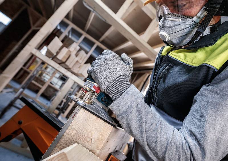 Ein Handwerker mit Schutzkleidung und Maske sägt ein Holzbrett in einer Werkstatt mit einer Kreissäge. Holzblöcke im Hintergrund.